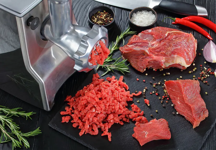 Metalowa maszynka do mielenia mięsa, na tacy leżą kawałki mięsa i przyprawy