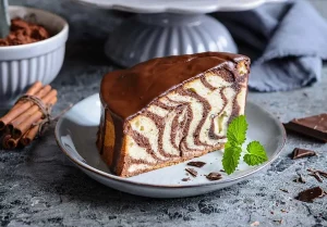 Przepis na ciasto zebra, ciasto zebra na jasnym talerzyku, w tle widać kakao, czekoladę oraz cynamon, ciasto udekorowane jest listkiem mięty.